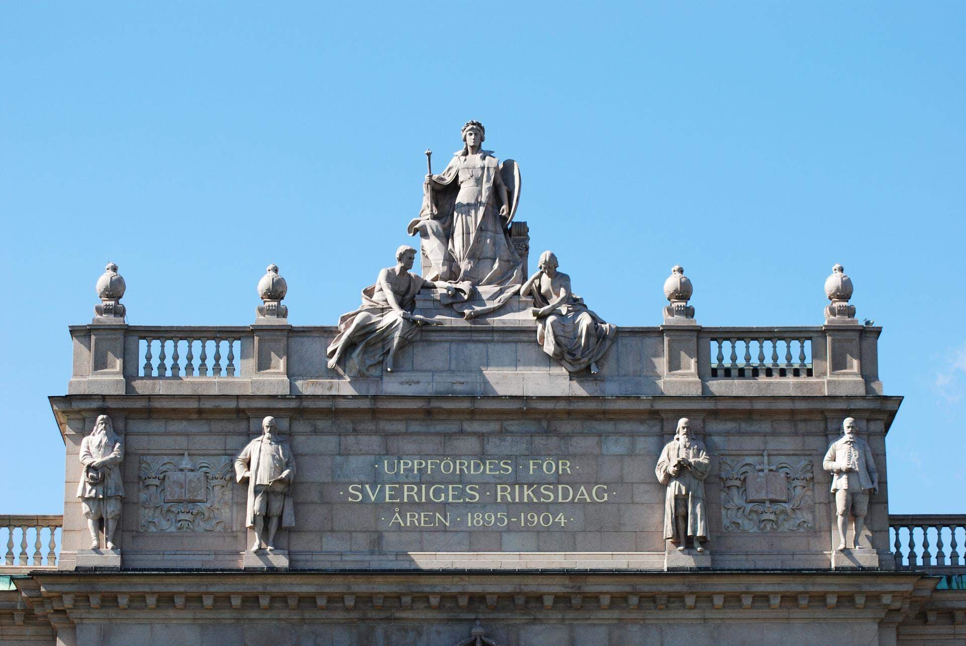 Statyerna i toppen på byggnaden för Sveriges Riksdag mot blå himmel